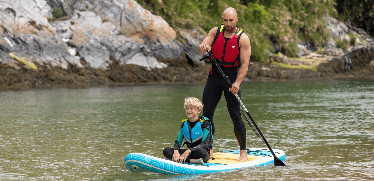 Man and child wearing PFDs while paddleboarding on calm water, demonstrating essential paddleboard safety practices.