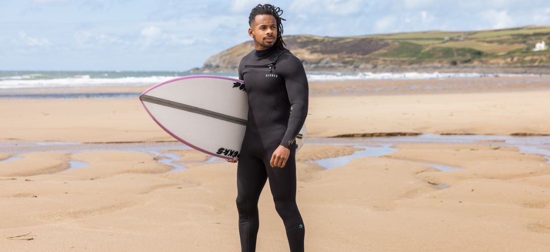 A man stood on a beach wearing a black Eco wetsuit. He is holding a white surfboard.