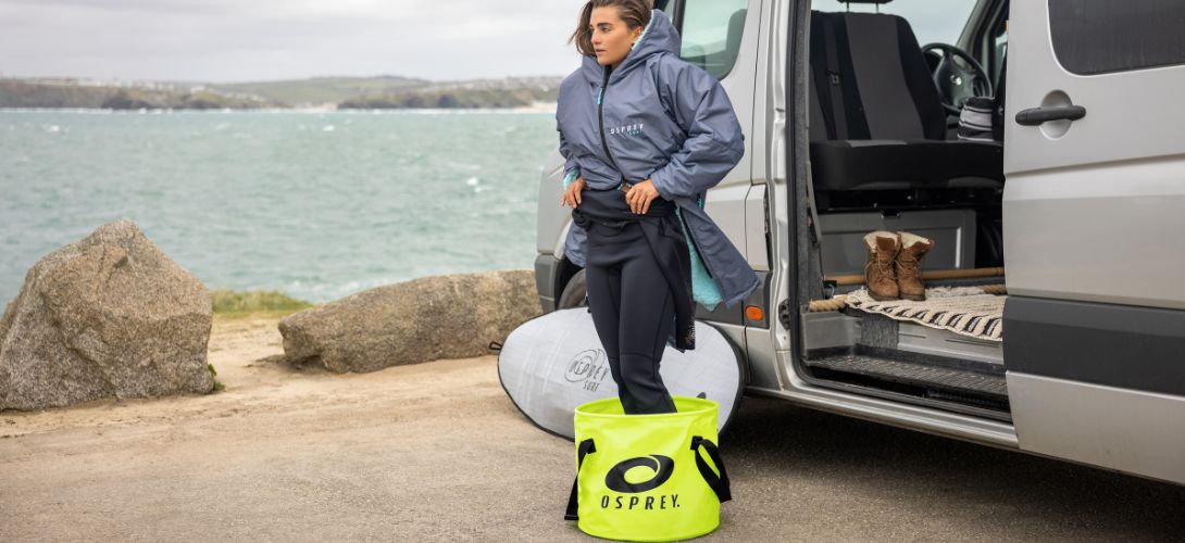A woman in a changing robe and wetsuit standing in a yellow Osprey changing bucket outside a van