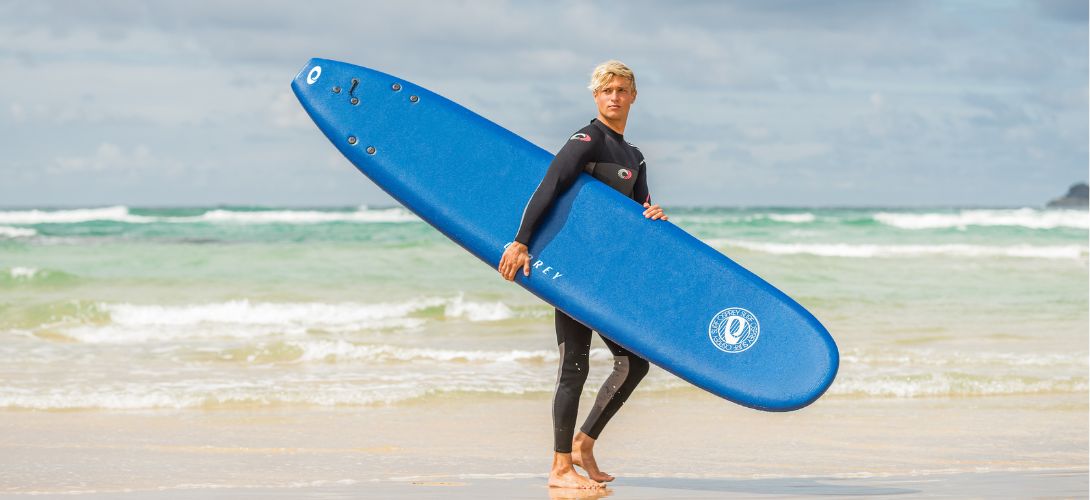 A photo of a man on the beach carrying a 9ft blue foam surfboard under his arm