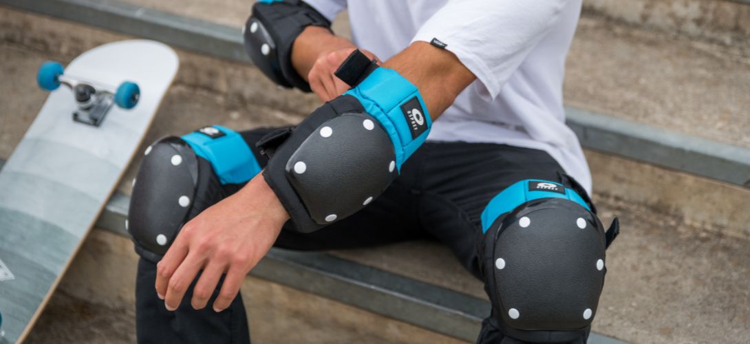 A close up photo of a man adjusting a set of Osprey elbow pads. He is sat down with a grey skateboard rested next to him