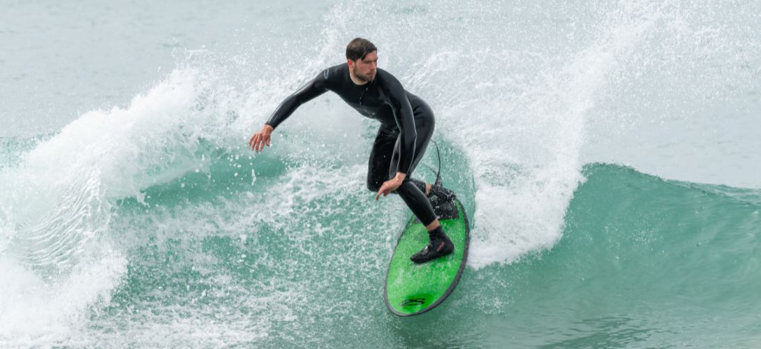 A man surfing a wave in a black wetsuit. He is on a green surfboard.