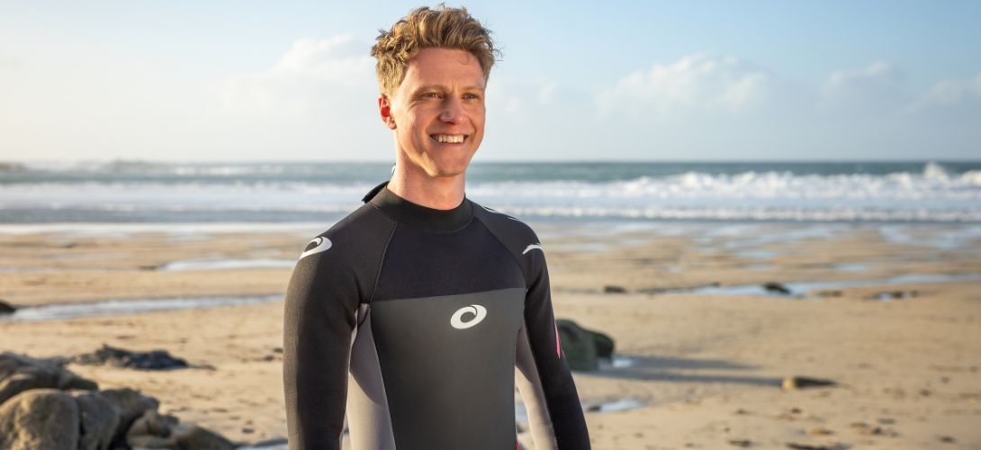 A close up of a man stood on a beach smiling wearing a black and red wetsuit