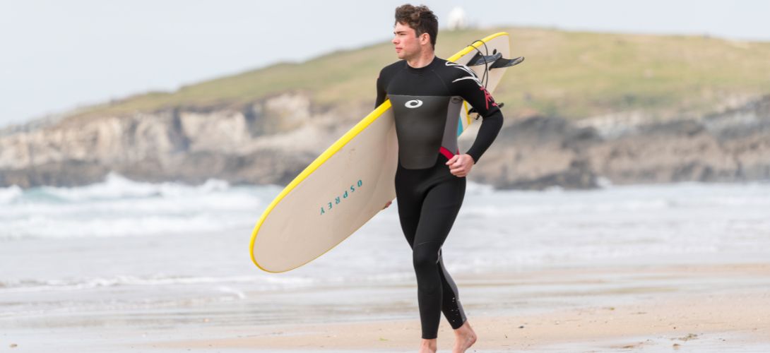A man running along a beach carrying a surfboard. He is wearing a black and red wetsuit