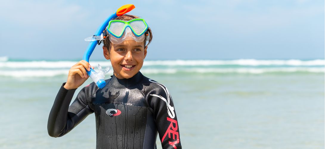 A photo of a boy stood in front of the sea, he is wearing a snorkel set with the goggles resting on his forehead