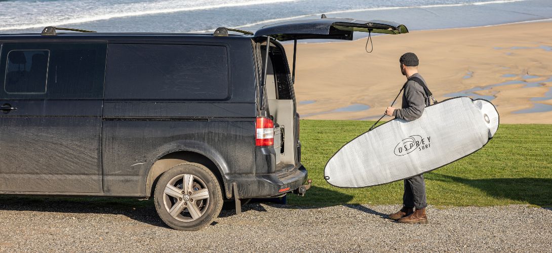 A man stood next to a black VW van. He is carrying a surfboard in a silver bag
