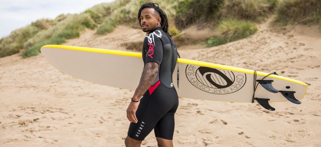 a man stood amongst sand dunes on a beach holding a yellow surfboard. He is a wearing a short black and red wetsuit.