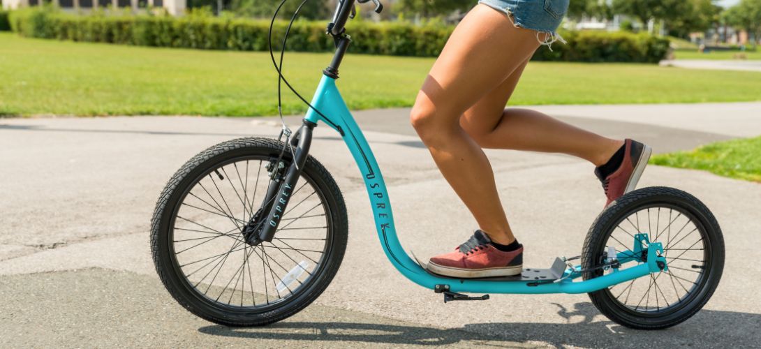 A close up photo of a woman riding a blue big wheeled scooter through a park
