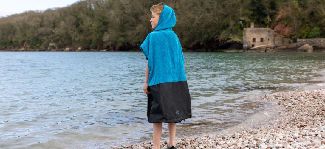 a young boy stood at a sea shore wearing a blue towel poncho