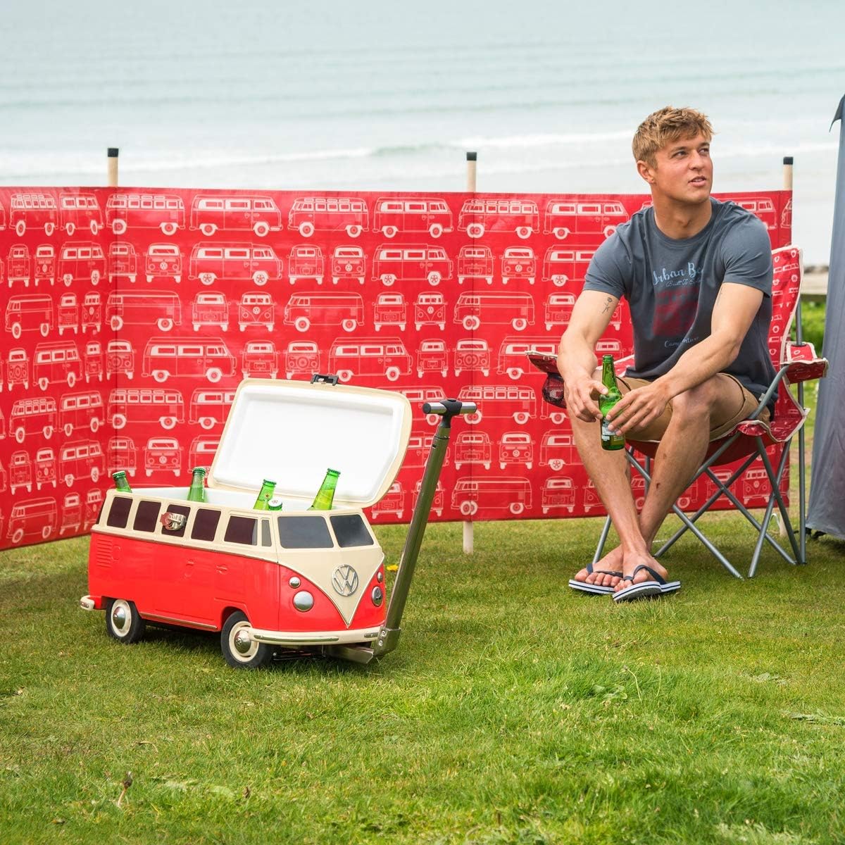 Person sitting on a chair next to a red and white Volkswagen camper van-shaped cooler with drinks on a grassy area.