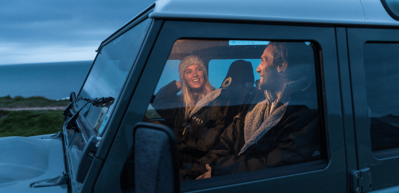 Two people wearing Osprey changing robes inside a Land Rover Defender, staying warm after cold-water surfing on a UK beach