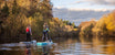 Two women paddleboarding down a river in autumn, the trees either side are orange and then sun is low
