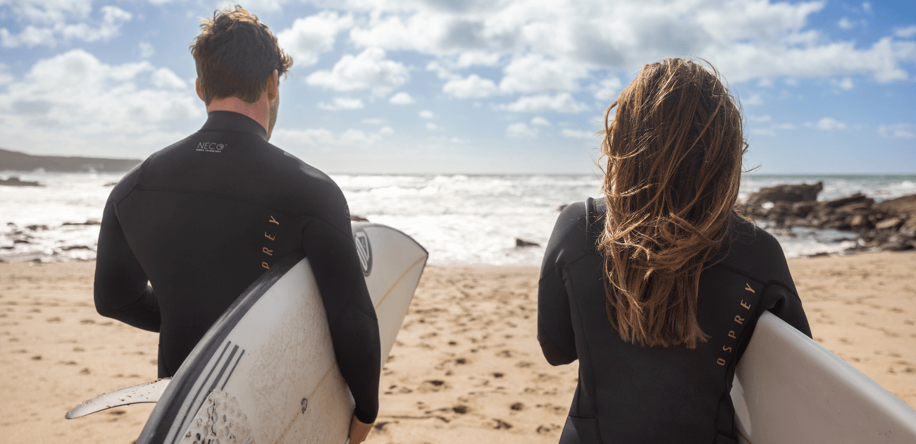Two surfers standing on the beach with their boards, representing the mental and physical wellbeing benefits of surfing and skating.