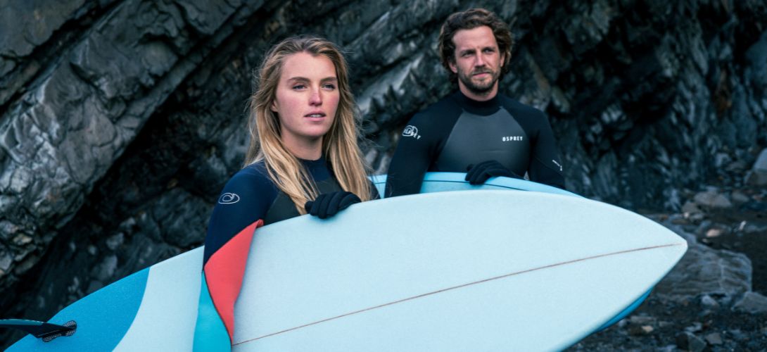 A photo of a man and woman against some black rocks. They are wearing wetsuits and then woman is holding a white surfboard