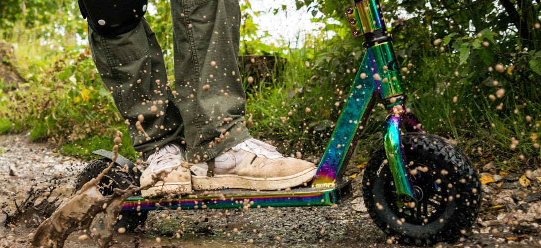 A close up of a neo-chrome dirt scooter being ridden through mud