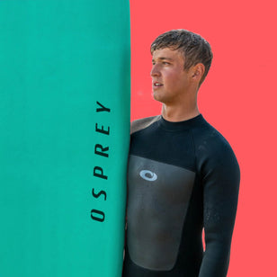 A male model stood next to a mint green Osprey foam surfboard against a coral background